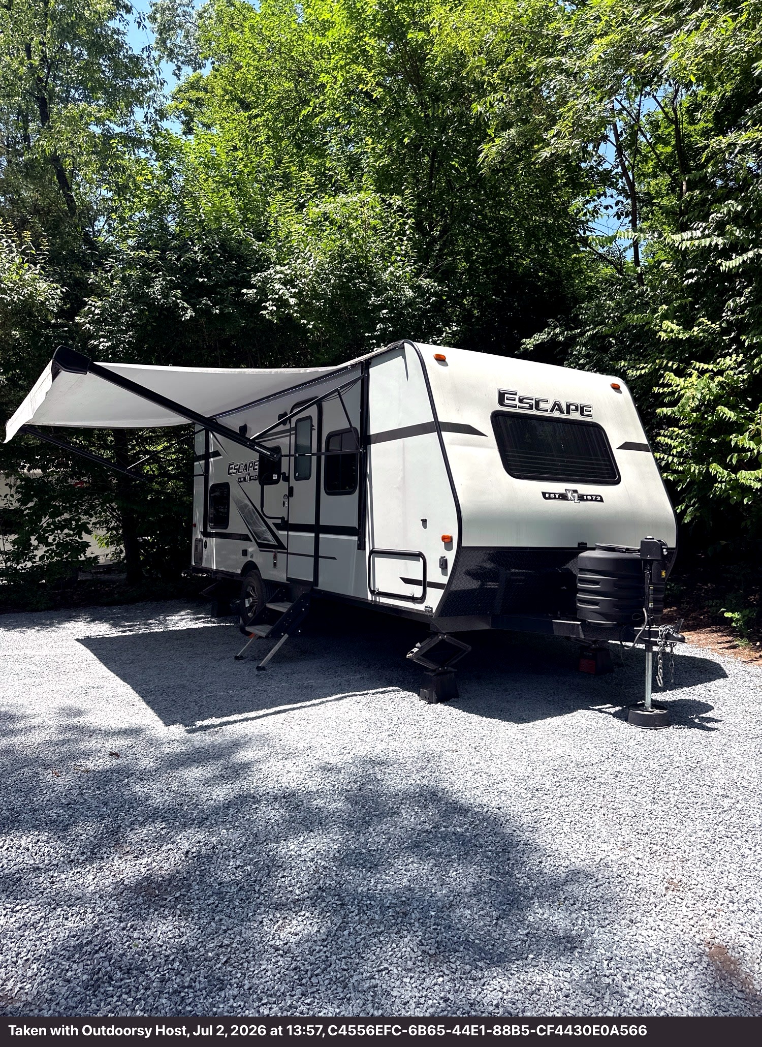 Escape travel trailer RV rental parked on gravel at a shaded campsite, surrounded by green trees, offering an outdoor escape.