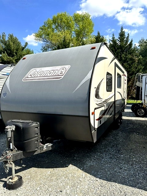 Coleman rv rental travel trailer, gray and beige, parked on gravel. Ready for camping trips and outdoor adventures.