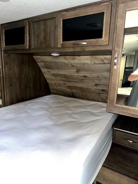 Inside an RV rental, this cozy bedroom features a comfortable bed, wood-look headboard, overhead cabinets, and mirrored nightstand.