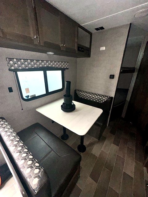 RV rental dinette area with dark patterned benches, white table, large window, and wood-toned overhead cabinets.