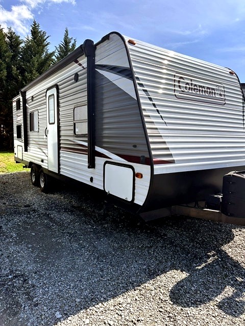 A sleek grey and white Coleman travel trailer, featuring an awning, ready for rv rental adventures on a gravel lot.