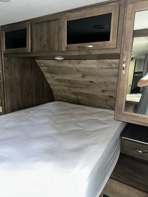 Inside an RV rental, this cozy bedroom features a comfortable bed, wood-look headboard, overhead cabinets, and mirrored nightstand.