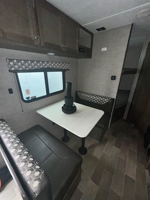 RV rental dinette area with dark patterned benches, white table, large window, and wood-toned overhead cabinets.